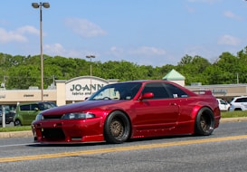 A bright red sports car with a sleek, modified body kit is parked on a road in front of a shopping center with various stores including Jo-Ann Fabrics and Crafts. The weather is sunny with a clear blue sky, and there are green trees in the background. Other vehicles are visible in the parking lot.