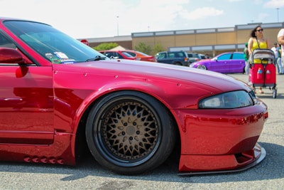 A bright red, heavily modified car is prominently displayed in a parking lot, featuring custom wheels and lowered suspension. In the background, a purple car is parked nearby, while people walk around, including a woman with a stroller.