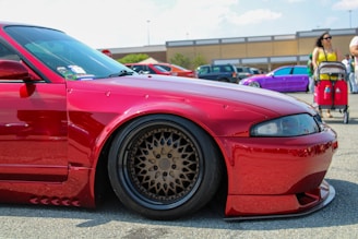 A bright red, heavily modified car is prominently displayed in a parking lot, featuring custom wheels and lowered suspension. In the background, a purple car is parked nearby, while people walk around, including a woman with a stroller.