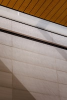 A wall surfaces composed of horizontal tiles with a light beige color dominates the lower portion of the image. The top portion features a section of yellow wood planks on a ceiling with black lines accentuating the divisions. A glass panel or glazed section with condensation is visible between the ceiling and wall.