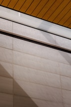 A wall surfaces composed of horizontal tiles with a light beige color dominates the lower portion of the image. The top portion features a section of yellow wood planks on a ceiling with black lines accentuating the divisions. A glass panel or glazed section with condensation is visible between the ceiling and wall.