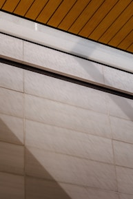 A wall surfaces composed of horizontal tiles with a light beige color dominates the lower portion of the image. The top portion features a section of yellow wood planks on a ceiling with black lines accentuating the divisions. A glass panel or glazed section with condensation is visible between the ceiling and wall.