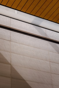 A wall surfaces composed of horizontal tiles with a light beige color dominates the lower portion of the image. The top portion features a section of yellow wood planks on a ceiling with black lines accentuating the divisions. A glass panel or glazed section with condensation is visible between the ceiling and wall.