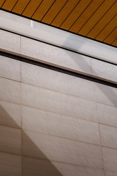 A wall surfaces composed of horizontal tiles with a light beige color dominates the lower portion of the image. The top portion features a section of yellow wood planks on a ceiling with black lines accentuating the divisions. A glass panel or glazed section with condensation is visible between the ceiling and wall.