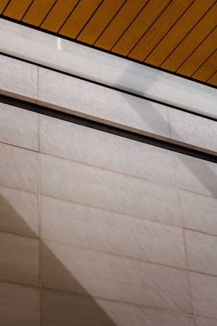 A wall surfaces composed of horizontal tiles with a light beige color dominates the lower portion of the image. The top portion features a section of yellow wood planks on a ceiling with black lines accentuating the divisions. A glass panel or glazed section with condensation is visible between the ceiling and wall.