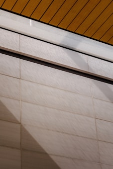 A wall surfaces composed of horizontal tiles with a light beige color dominates the lower portion of the image. The top portion features a section of yellow wood planks on a ceiling with black lines accentuating the divisions. A glass panel or glazed section with condensation is visible between the ceiling and wall.