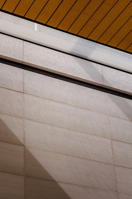 A wall surfaces composed of horizontal tiles with a light beige color dominates the lower portion of the image. The top portion features a section of yellow wood planks on a ceiling with black lines accentuating the divisions. A glass panel or glazed section with condensation is visible between the ceiling and wall.