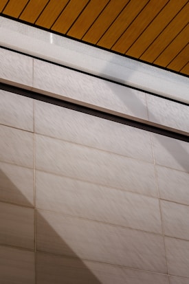 A wall surfaces composed of horizontal tiles with a light beige color dominates the lower portion of the image. The top portion features a section of yellow wood planks on a ceiling with black lines accentuating the divisions. A glass panel or glazed section with condensation is visible between the ceiling and wall.