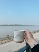 A hand holding a white mug with the logo 'HONEY MOON COFFEE CO' is held up against a scenic backdrop of a river and a clear blue sky. The scene includes a railing and a road lined with young trees, conveying a calm and peaceful setting.