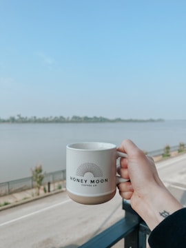 A hand holding a white mug with the logo 'HONEY MOON COFFEE CO' is held up against a scenic backdrop of a river and a clear blue sky. The scene includes a railing and a road lined with young trees, conveying a calm and peaceful setting.