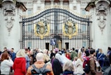 A group of people gathered in front of ornate, gold-adorned black iron gates with a large emblem. Some individuals are holding British flags and wearing patriotic clothing. The gates feature detailed stone carvings and are part of an imposing classical-style building.