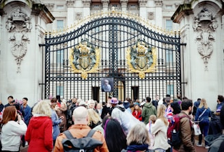 A group of people gathered in front of ornate, gold-adorned black iron gates with a large emblem. Some individuals are holding British flags and wearing patriotic clothing. The gates feature detailed stone carvings and are part of an imposing classical-style building.