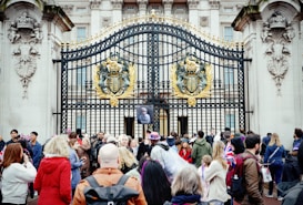 A group of people gathered in front of ornate, gold-adorned black iron gates with a large emblem. Some individuals are holding British flags and wearing patriotic clothing. The gates feature detailed stone carvings and are part of an imposing classical-style building.
