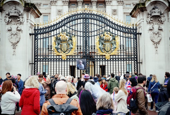 A group of people gathered in front of ornate, gold-adorned black iron gates with a large emblem. Some individuals are holding British flags and wearing patriotic clothing. The gates feature detailed stone carvings and are part of an imposing classical-style building.