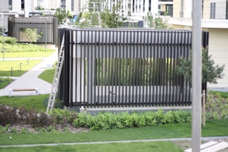 A modern outdoor structure with vertical slats, situated in a well-maintained garden area. There is a ladder leaning against the structure, which is surrounded by grass and pathways. Trees and shrubs are interspersed throughout the landscaped area, and there are benches and lighting fixtures along the pathways. Buildings in the background suggest an urban setting.