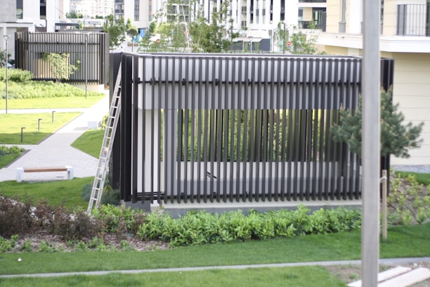 A modern outdoor structure with vertical slats, situated in a well-maintained garden area. There is a ladder leaning against the structure, which is surrounded by grass and pathways. Trees and shrubs are interspersed throughout the landscaped area, and there are benches and lighting fixtures along the pathways. Buildings in the background suggest an urban setting.