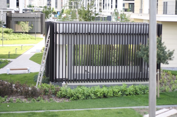 A modern outdoor structure with vertical slats, situated in a well-maintained garden area. There is a ladder leaning against the structure, which is surrounded by grass and pathways. Trees and shrubs are interspersed throughout the landscaped area, and there are benches and lighting fixtures along the pathways. Buildings in the background suggest an urban setting.