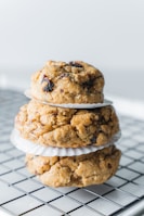 Three large, round cookies with a rustic texture are stacked on top of each other with paper liners separating each cookie. The cookies contain visible chunks of what appears to be dried fruit or chocolate chips. The stack is placed on a cooling rack.