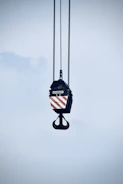 A large industrial crane hook is suspended in the air, hanging from several thick cables. The hook block features a pattern of diagonal red and white stripes. The background is a pale blue sky.
