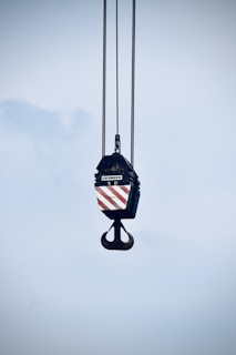 A large industrial crane hook is suspended in the air, hanging from several thick cables. The hook block features a pattern of diagonal red and white stripes. The background is a pale blue sky.