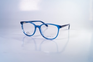 A pair of stylish, blue-rimmed glasses with transparent lenses is placed on a reflective white surface. The brand name 'Esprit' is printed on one of the lenses, along with the words 'Small collection'. The glasses' design is modern and sleek, with a lightweight frame.