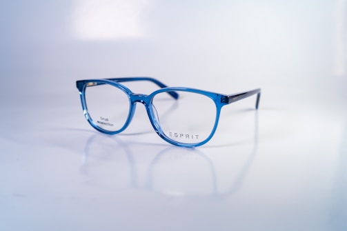 A pair of stylish, blue-rimmed glasses with transparent lenses is placed on a reflective white surface. The brand name 'Esprit' is printed on one of the lenses, along with the words 'Small collection'. The glasses' design is modern and sleek, with a lightweight frame.