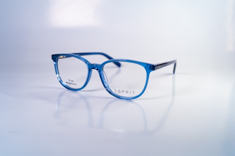 A pair of stylish, blue-rimmed glasses with transparent lenses is placed on a reflective white surface. The brand name 'Esprit' is printed on one of the lenses, along with the words 'Small collection'. The glasses' design is modern and sleek, with a lightweight frame.