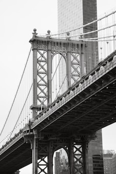 A close-up perspective of a suspension bridge with intricate metalwork. The frame captures a prominent bridge tower and the suspension cables leading to the roadbed. In the background, a modern glass skyscraper is partially visible, complementing the bridge's classic architecture. The scene is in black and white, emphasizing the design and structure details.