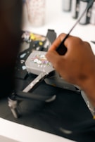 A person's hand is painting intricate floral designs on a gray drone. The background is blurred, with art supplies visible, including brushes and paint bottles.