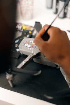 A person's hand is painting intricate floral designs on a gray drone. The background is blurred, with art supplies visible, including brushes and paint bottles.