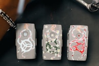 Three gray drone batteries are artistically decorated with colorful designs. The first battery features white swirling patterns with floral and dynamic motifs. The second battery showcases floral designs in green and white, forming intricate patterns. The third battery is adorned with bold red and white designs, including heart shapes.