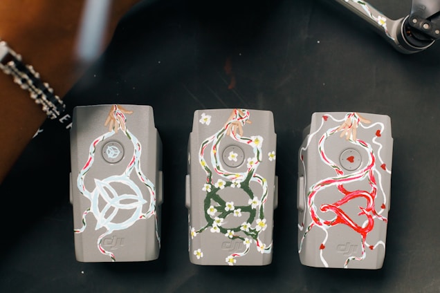 Three gray drone batteries are artistically decorated with colorful designs. The first battery features white swirling patterns with floral and dynamic motifs. The second battery showcases floral designs in green and white, forming intricate patterns. The third battery is adorned with bold red and white designs, including heart shapes.