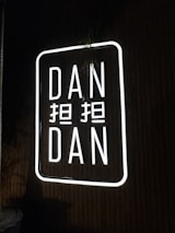 A neon sign displaying the words 'DAN DAN' with additional characters between them. The sign is illuminated with bright white light against a dark background.