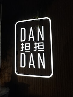 A neon sign displaying the words 'DAN DAN' with additional characters between them. The sign is illuminated with bright white light against a dark background.