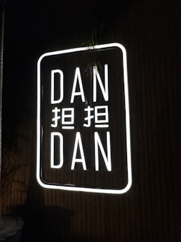 A neon sign displaying the words 'DAN DAN' with additional characters between them. The sign is illuminated with bright white light against a dark background.