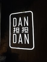 A neon sign displaying the words 'DAN DAN' with additional characters between them. The sign is illuminated with bright white light against a dark background.