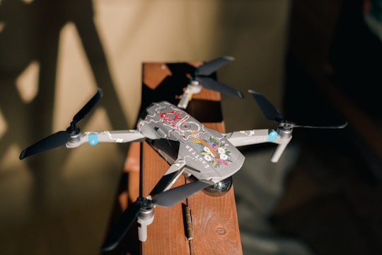 A drone with four black propellers rests on a wooden surface, illuminated by sunlight. The drone's body features colorful decals with floral patterns, adding a decorative touch to its sleek design. The shadows on the background enhance the composition, creating a warm and inviting atmosphere.