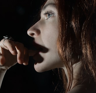 A close-up profile of a person with fair skin and red hair. The individual is gazing thoughtfully into the distance with a finger near their lips. The lighting is dim, creating a dramatic contrast and highlighting their facial features.