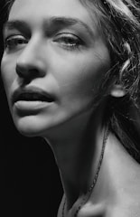 A monochrome portrait of a person with prominent facial features, including detailed eyes and textured skin. The lighting is dramatic, with strong contrasts highlighting the contours of the face and neck.