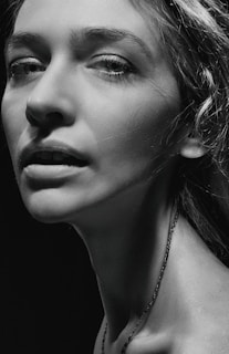 A monochrome portrait of a person with prominent facial features, including detailed eyes and textured skin. The lighting is dramatic, with strong contrasts highlighting the contours of the face and neck.