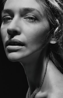 A monochrome portrait of a person with prominent facial features, including detailed eyes and textured skin. The lighting is dramatic, with strong contrasts highlighting the contours of the face and neck.