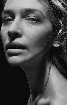 A monochrome portrait of a person with prominent facial features, including detailed eyes and textured skin. The lighting is dramatic, with strong contrasts highlighting the contours of the face and neck.