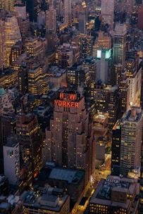 Aerial view of a densely packed urban landscape with towering skyscrapers illuminated by numerous lights. The prominent building features a sign that reads 'NEW YORKER'. Tall structures, diverse architectural designs, and a maze of streets are visible.