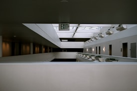 A modern indoor architectural space featuring multiple levels with an open design. Each level has a series of doors along the hallway, illuminated by wall-mounted lights. Potted plants are aligned on ledges, adding greenery to the sterile environment. A prominent 'EXIT' sign hangs from the ceiling, indicating the direction for an emergency exit. The overall aesthetic is minimalistic with clean lines and neutral tones.