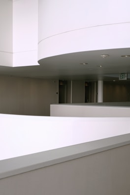 A minimalist interior hallway with smooth, curved architectural lines. The walls and ceiling are predominantly white, providing a clean and modern aesthetic. Recessed lighting fixtures are embedded in the ceiling, and there is an exit sign visible in the background.