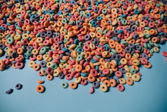A large amount of colorful, circular cereal pieces spread out on a light blue surface. The cereal pieces are in various colors, including red, green, yellow, orange, purple, and blue.