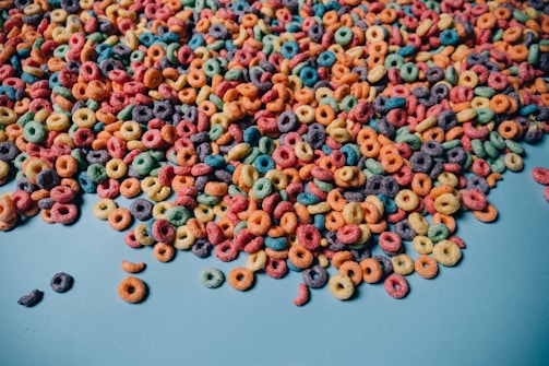 A large amount of colorful, circular cereal pieces spread out on a light blue surface. The cereal pieces are in various colors, including red, green, yellow, orange, purple, and blue.