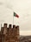 A stone castle wall towers in the foreground with the Portuguese flag flying high on a flagpole. The sky is overcast with a light, cloudy atmosphere, giving a muted tone to the scene. In the background, a vast cityscape stretches out, with numerous buildings and a distant bridge.