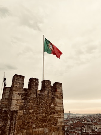 A stone castle wall towers in the foreground with the Portuguese flag flying high on a flagpole. The sky is overcast with a light, cloudy atmosphere, giving a muted tone to the scene. In the background, a vast cityscape stretches out, with numerous buildings and a distant bridge.
