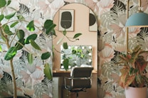 A room with tropical botanical wallpaper featuring large leaves and floral patterns. A decorative mirror with lights is positioned above a wooden desk, accompanied by a comfortable swivel chair. Plants, both potted and in the wallpaper design, add a lush, vibrant feel to the space. Artwork hangs on the wall, and a modern lamp with a blue shade contributes to the eclectic decor.